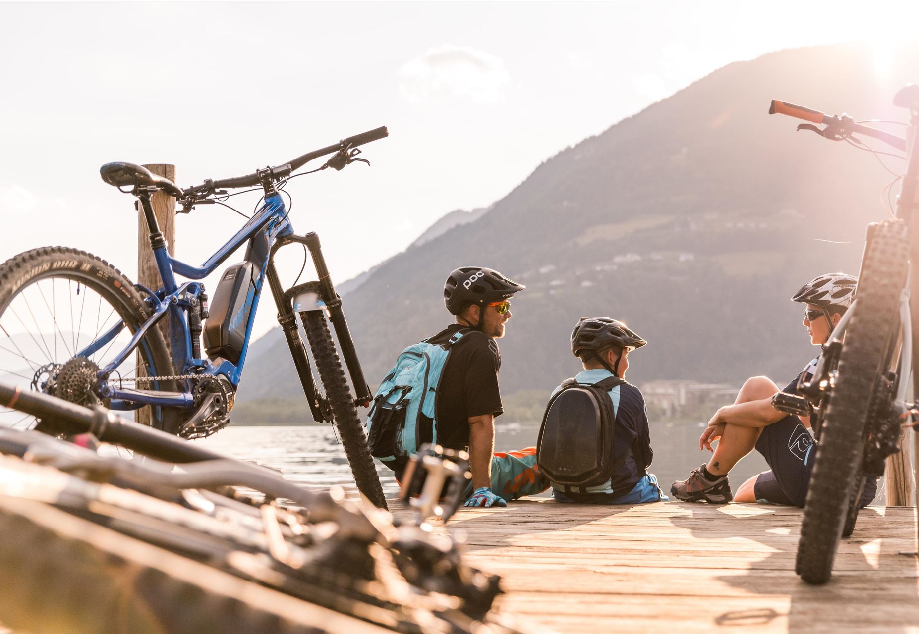 Drei Kinder sitzen am Ufer eines Sees auf einem Holzsteg. Im Vordergrund stehen Mountainbikes und im Hintergrund sind Berge zu sehen.