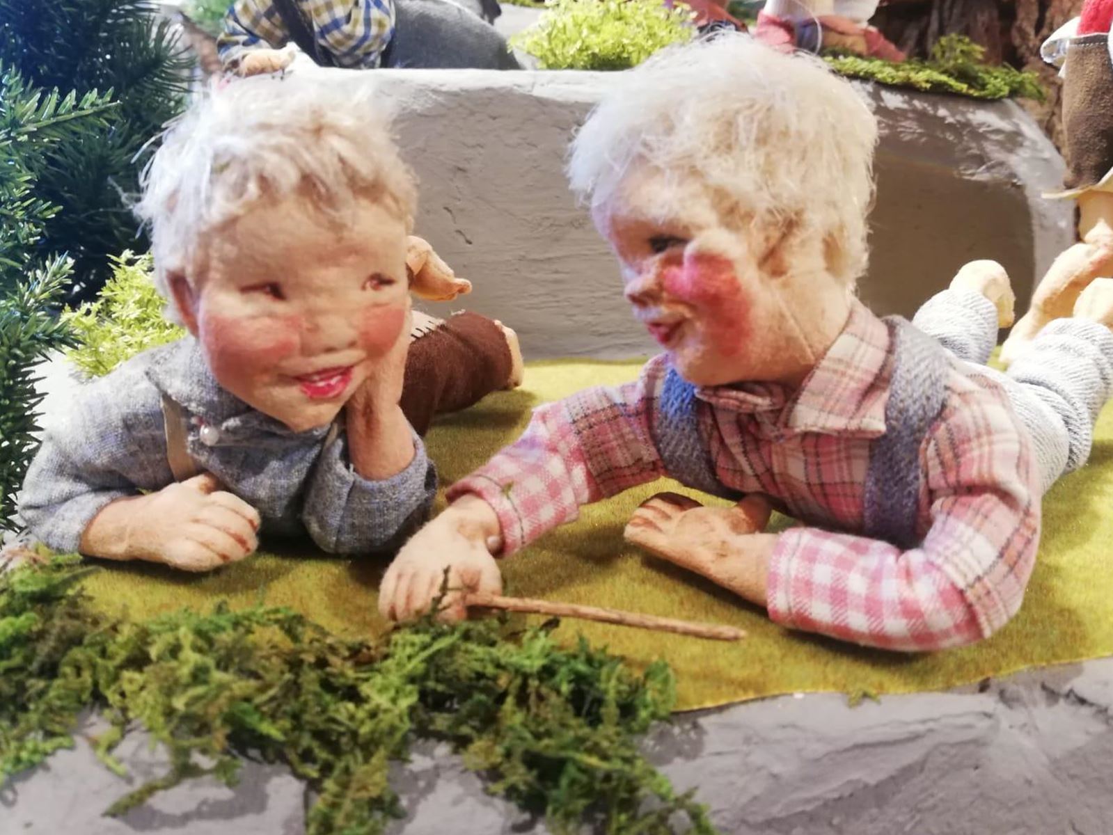 Zwei Keramikkinder spielen fröhlich auf einer Wiese. Sie liegen nebeneinander und scheinen einander etwas zu erzählen.