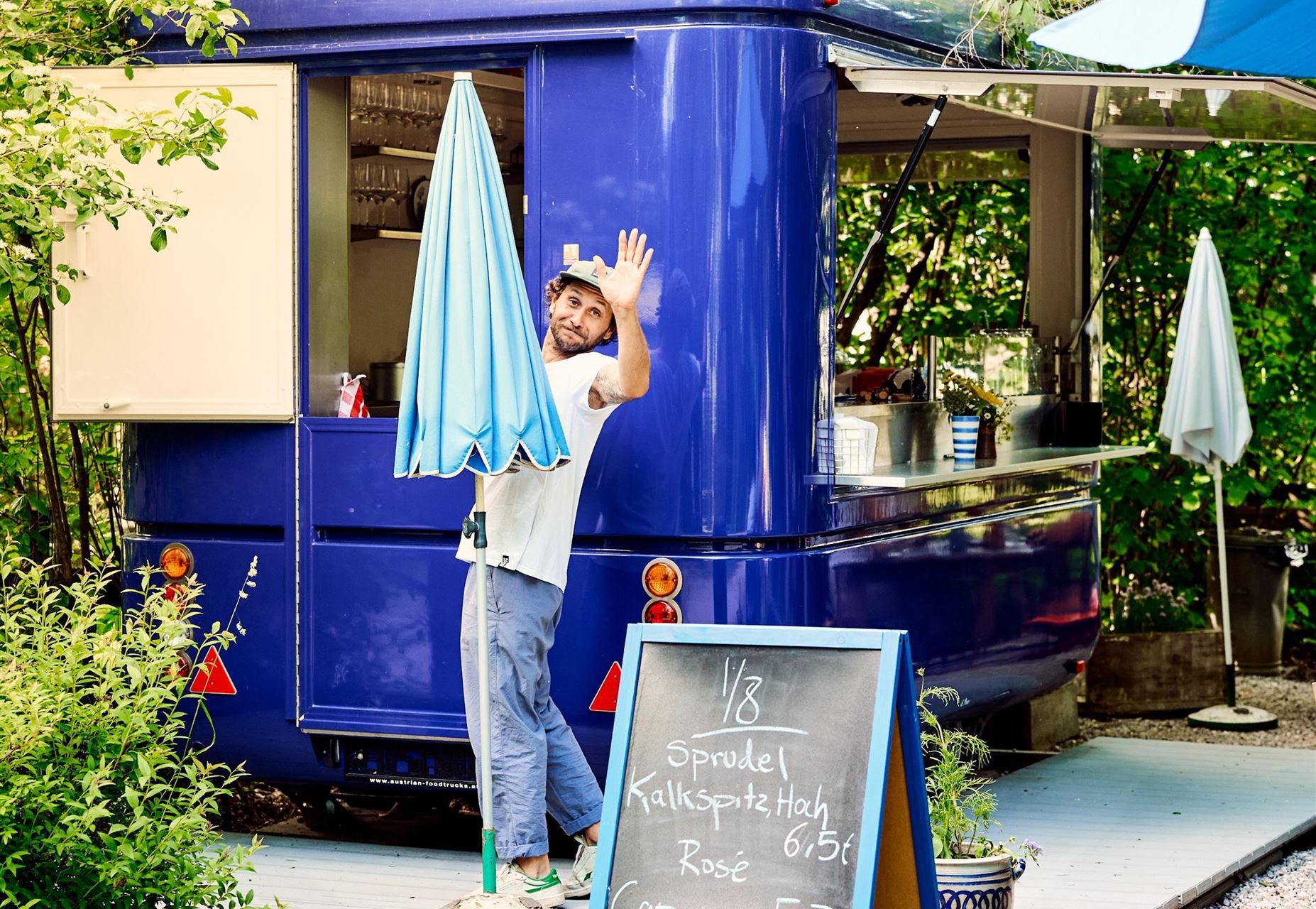 Ein gemütlicher Foodtruck in der Natur, umgeben von Bäumen. Ein freundlicher Mann winkt vor dem Truck, während ein Schirm aufgestellt wird.