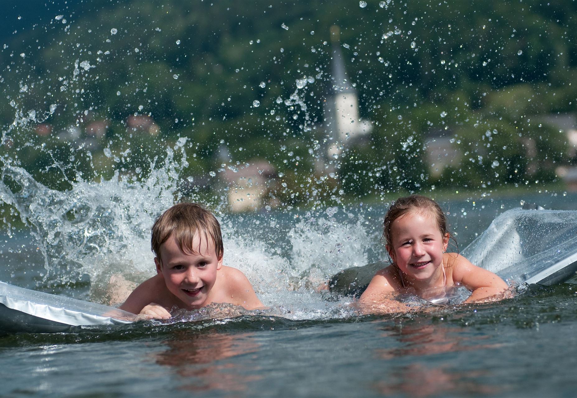 Zwei Kinder schwimmen fröhlich im Wasser und spritzen dabei. Im Hintergrund ist eine malerische Landschaft mit einem Kirchturm zu sehen.