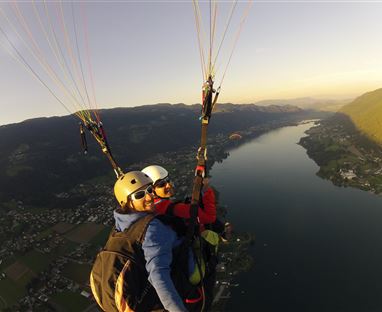Zwei Paraglider fliegen hoch über einem klaren See und einer wunderschönen Landschaft. Die Sonne strahlt am Himmel und schafft eine malerische Aussicht.