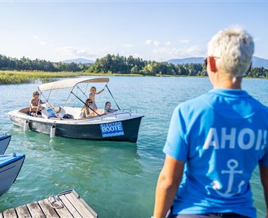 Ein Boot mit mehreren Personen fährt auf einem blauen See. Im Vordergrund steht eine Person mit einem blauen T-Shirt, das "AHOI!" zeigt.
