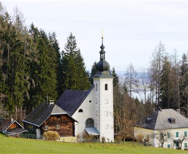 Eine malerische Kirche mit einem hohen Turm, umgeben von grünen Wäldern. Im Hintergrund sind sanfte Hügel und Bäume sichtbar.