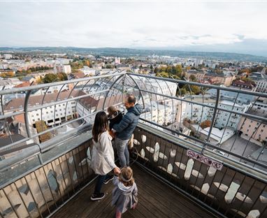 Eine Aussichtspunkt mit einem Blick auf die Stadt. Drei Personen genießen die Aussicht auf die umliegenden Gebäude und Landschaft.