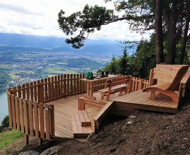 Eine Holzterrasse mit Blick auf einen See und die umliegende Landschaft. Die Terrasse verfügt über Sitzgelegenheiten und liegt idyllisch am Hang.