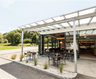Ein modernes Café mit Außensitzplätzen und großer Fensterfront. Umgeben von einer grünen Landschaft und einem klaren Himmel.