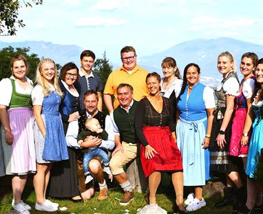 Eine Gruppe von Menschen in traditioneller Tracht steht vor einer malerischen Landschaft. Im Hintergrund sind Berge und ein klarer Himmel zu sehen.