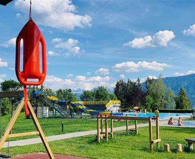 Ein schöner Park mit einem Schwimmbad und einer Rutsche im Hintergrund. Im Vordergrund hängt eine Rettungsboje über einer Schaukel.