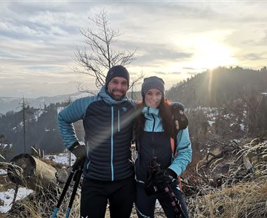 Una coppia si trova in un paesaggio invernale con attrezzatura da sci. Sullo sfondo si possono vedere montagne coperte di neve e un'alba.