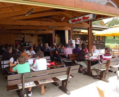 A fully occupied restaurant with many guests sitting at long wooden tables. The atmosphere is cozy and inviting.