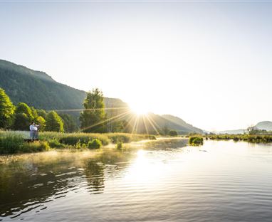 A picturesque river with gentle hills in the background. The sun is rising and creating a bright, warm atmosphere.