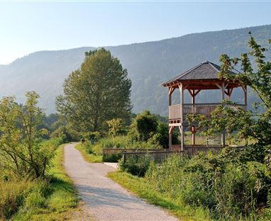 Ein malerischer Weg führt zu einem Aussichtspunkt mit einem überdachten Pavillon. Im Hintergrund sind sanfte Hügel und Bäume zu sehen.