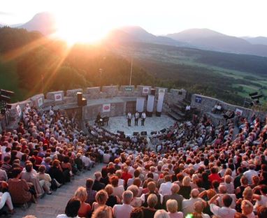 Ein großes Publikum sitzt in einem Freilufttheater, umgeben von Bergen. Die Sonne geht hinter den Gipfeln unter und taucht die Szene in warmes Licht.