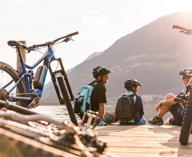 Drei Kinder sitzen am Ufer eines Sees auf einem Holzsteg. Im Vordergrund stehen Mountainbikes und im Hintergrund sind Berge zu sehen.