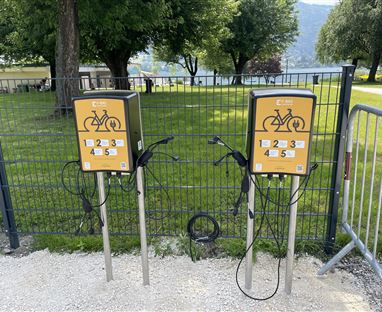 Zwei Fahrradladestationen stehen an einem Zaun in einem Park. Die Stationen sind gelb mit vorgesehenen Plätzen für Fahrräder.
