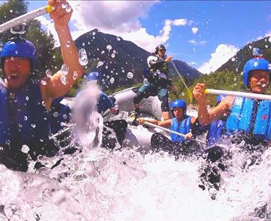 Eine Gruppe von Menschen macht Wildwassertour im Raftingboot. Sie tragen Schwimmwesten und Helme und haben viel Spaß im spritzenden Wasser.
