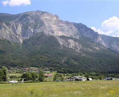 Eine weite Landschaft mit Bergen im Hintergrund und einer Wiese im Vordergrund. Im Bild sind auch einige Flugzeuge und Gebäude zu sehen.