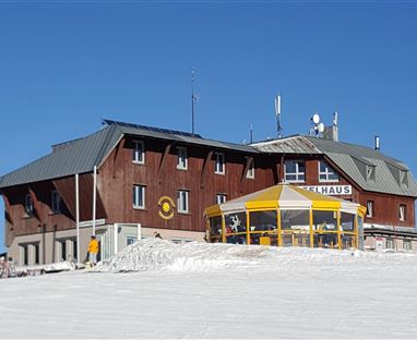 A mountain house with a wooden facade in a snow-covered area. The sky is clear and blue, and some skiers are nearby.