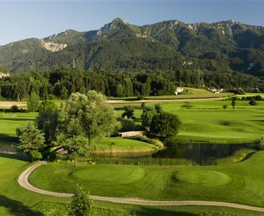 Eine Landschaft mit einem Golfplatz, umgeben von sanften Hügeln und Bäumen. Im Hintergrund sind majestätische Berge zu sehen.