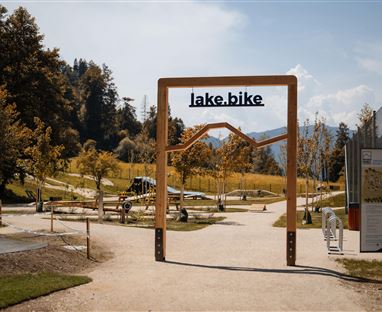 Un cartello di accesso per lake.bike in un ambiente paesaggistico. Il sentiero attraversa un parco con alberi e opportunità ricreative.