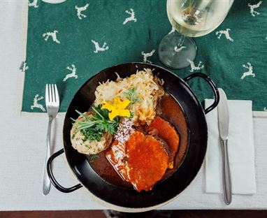 A delicious dish on a black plate, garnished with fresh herbs and a flower. There is also a glass plate with a drink and cutlery on a green table runner.