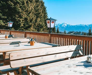A cozy outdoor area with wooden tables and benches. In the background, majestic mountains rise under a clear blue sky.