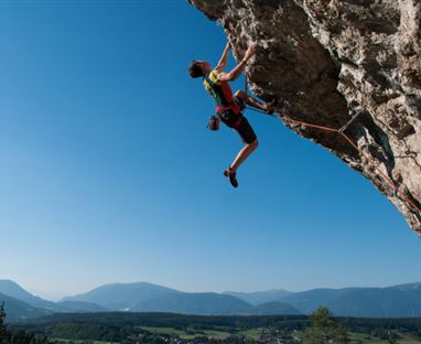 Ein Kletterer in einem roten Klettergeschirr, der an einer Felswand hochklettert. Im Hintergrund sind Berge und ein klarer blauer Himmel zu sehen.