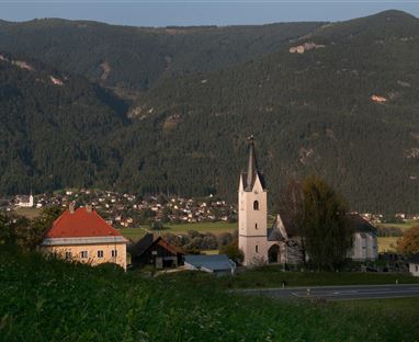 Eine malerische Landschaft mit einem kleinen Dorf und einer Kirche im Vordergrund. Im Hintergrund sind sanfte Berge und Wälder zu sehen.