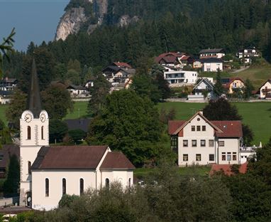 Ein malerisches Dorf mit einer weißen Kirche und modernen Häusern. Umgeben von grünen Bergen und Wäldern.