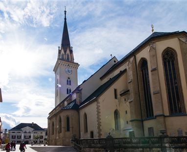 Eine historische Kirche mit einem hohen Turm in einer Stadt. Das Licht der Sonne strahlt durch den Himmel und beleuchtet die Gebäude in der Umgebung.