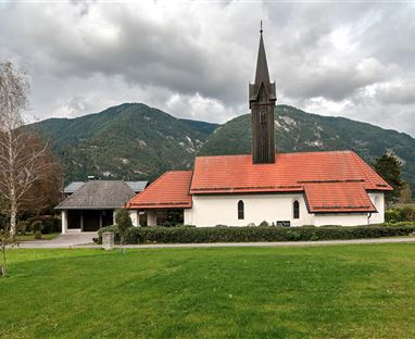 Eine kleine Kirche mit einem roten Dach steht auf einer Wiese. Im Hintergrund sind grüne Berge und ein bewölkter Himmel zu sehen.