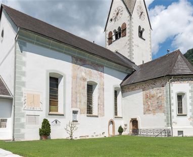 Eine alte Kirche mit weißen Wänden und bunten Fresken. Der grüne Rasen und die umliegenden Berge schaffen eine malerische Kulisse.