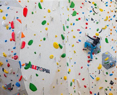Un climber sta scalando una colorata parete di bouldering in una palestra di arrampicata. La parete è dotata di molti prese diverse in vari colori.