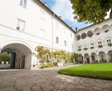 Ein schöner Innenhof mit weißem Gebäude, grüner Rasenfläche und blühenden Pflanzen. Die Architektur ist klassisch und einladend.