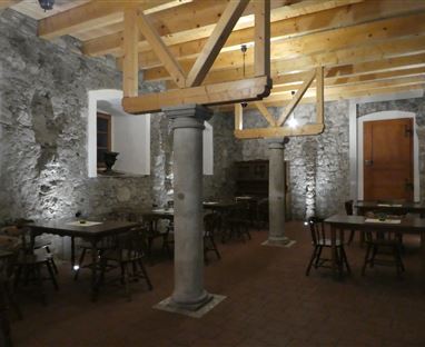 Ein gemütlicher Raum mit Steinwänden und Holzbalkendecke. Es gibt mehrere Tische und Stühle, die auf den Boden angeordnet sind.