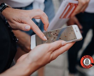 Una mano tiene uno smartphone, su una vista della mappa è visualizzata. Sullo sfondo si vedono persone e un opuscolo informativo.