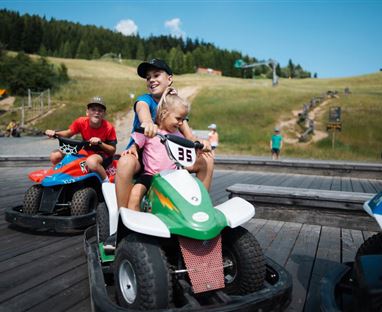 I bambini guidano i go-kart su una pista di legno nella natura. Sullo sfondo si vedono colline verdi e alberi.