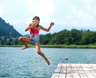 Ein Mädchen springt fröhlich von einem Steg ins Wasser. Im Hintergrund sind grüne Bäume und Berge zu sehen.