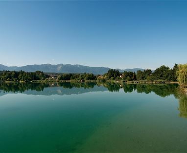 Ein ruhiger See mit klarem, grünem Wasser und einer malerischen Berglandschaft im Hintergrund. Der Himmel ist strahlend blau und die Umgebung ist von Bäumen gesäumt.