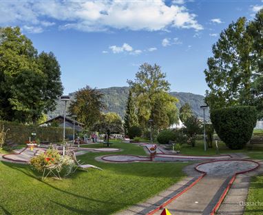 Eine Minigolfanlage mit gepflegten Grünflächen und Bäumen. Im Hintergrund sind sanfte Hügel und ein blauer Himmel zu sehen.