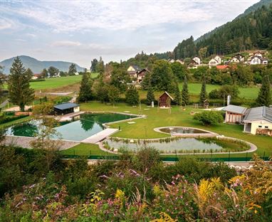 Eine idyllische Landschaft mit einem Schwimmteich und gepflegten Grünflächen. Im Hintergrund sind typische Wohnhäuser und sanfte Hügel zu sehen.