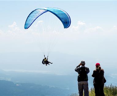 Ein Gleitschirmflieger schwebt über einer Berglandschaft. Im Vordergrund beobachten zwei Personen das Flugmanöver.
