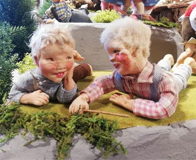 Zwei Keramikkinder spielen fröhlich auf einer Wiese. Sie liegen nebeneinander und scheinen einander etwas zu erzählen.