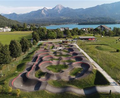Ein moderner Pumptrack in einer grünen Landschaft mit Bergen im Hintergrund. Der See glitzert in der Sonne, während Menschen aktiv auf dem Track sind.