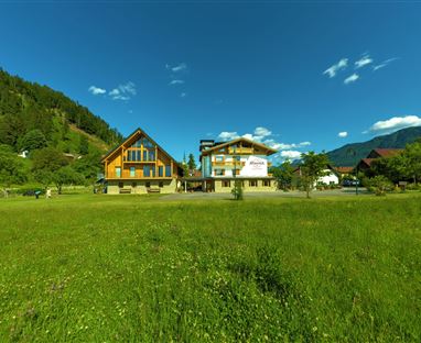 Eine malerische Landschaft mit zwei gemütlichen Gebäuden inmitten von grünem Gras. Der klare blaue Himmel und die umgebenden Hügel schaffen eine entspannte Atmosphäre.