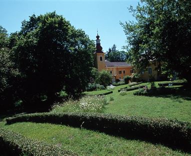 Eine idyllische Landschaft mit gepflegten Gärten und Bäumen. Im Hintergrund ist ein historisches Gebäude mit einem Turm zu sehen.