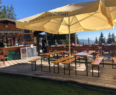 Eine rustikale Terrasse mit Holzbänken und Tischen unter einem großen Sonnenschirm. Im Hintergrund steht eine kleine Hütte in einer grünen Umgebung.
