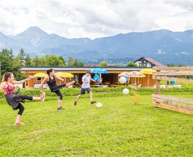 Drei Personen spielen im Freien mit einem Ball vor einer malerischen Berglandschaft. Im Hintergrund sind bunte Sonnenschirme und eine Holzkonstruktion zu sehen.