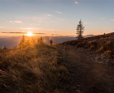 Ein Wanderer läuft auf einem Pfad in der Berglandschaft. Die Sonne geht hinter den Bergen unter und erleuchtet den Himmel.