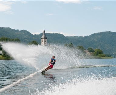 Ein Wasserski-Fahrer gleitet über einen ruhigen See. Im Hintergrund sind sanfte Hügel und ein hoher Kirchturm zu sehen.